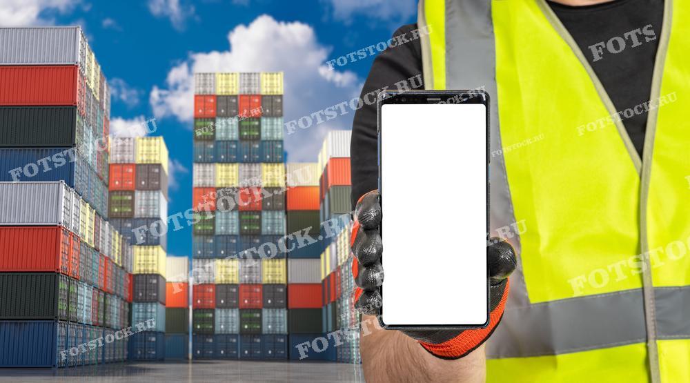 На изображении показан рабочий в защитном жилете, держащий смартфон с пустым экраном, на фоне многочисленных складированных контейнеров. Фотография иллюстрирует использование мобильных технологий и цифровых решений в логистике и управлении цепочками поставок, подчеркивая важность цифровизации в современной индустрии.